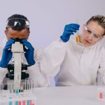 Dois cientistas de jaleco realizam pesquisa em laboratório, usando um microscópio e manipulando tubos de ensaio.